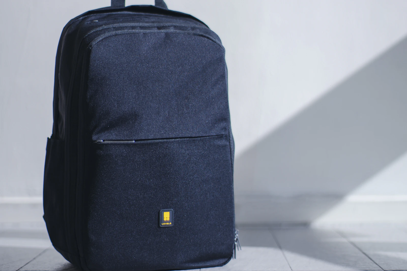 Daypack hitam berdiri di depan dinding terang dengan bayangan panjang